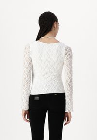 Weißes Spitzentop mit langen Ärmeln und tailliertem Schnitt. Verfügt über ein strukturiertes Blumenmuster und einen runden Ausschnitt. Kombiniert mit schwarzen Hosen.