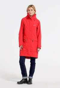 Veste imperméable rouge avec capuche, dotée de poches zippées et d'un design long. Associée à un jean foncé et des bottines noires.