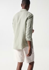 Camisa verde claro de manga larga con puños enrollados, con un diseño de dobladillo curvado. La prenda se lleva con pantalones cortos blancos hasta la rodilla. Tejido suave y texturizado.