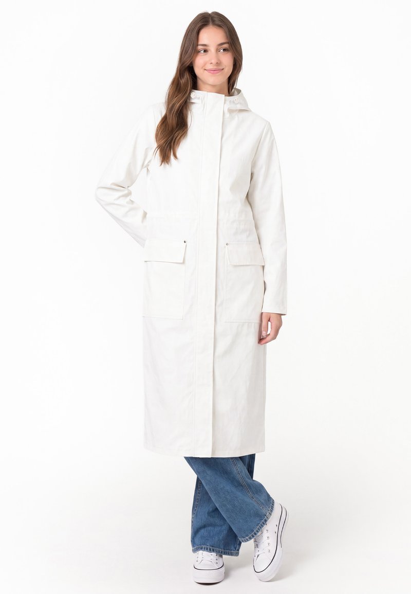 Jeune femme portant un long manteau blanc à capuche avec poches avant sur un jean bleu et des baskets blanches, debout les jambes croisées.