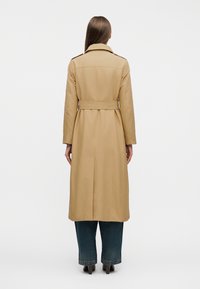 Γυναίκα με μακριά ίσια μαλλιά που φοράει ένα μπεζ παλτό τύπου τρανς με ζώνη, μπλε τζιν και παπούτσια με τακούνι, στέκεται γυρισμένη προς τα πίσω σε λευκό φόντο.