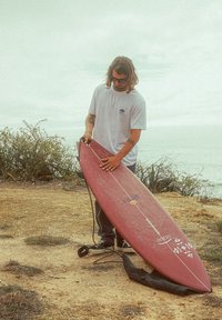 Planche de surf rouge avec des accents blancs, en matériau en fibre de verre, nez pointu, tenue par une personne en chemise blanche, se tenant sur un terrain sablonneux.