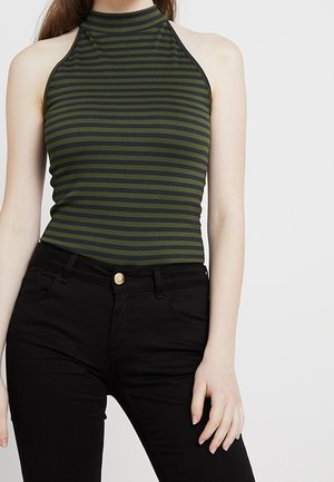 Kobieta w bezrękawnym zielono-czarnym topie w poziome paski i wysokich czarnych spodniach, stojąca na tle jednolitego tła.