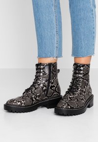 Bottines à imprimé serpent noir et gris avec une semelle en caoutchouc robuste, lacets sur le devant et fermeture éclair décorative sur le côté. Motif texturé effet peau de serpent.