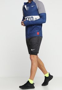 Man wearing long-sleeve navy and gray Nike top, black Nike shorts, neon yellow socks, and black Nike running shoes walking.