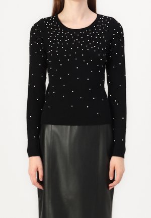 Femme portant un pull noir à manches longues avec des perles blanches éparses et une jupe en cuir noire, debout devant un fond uni.