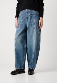 Vaqueros de mezclilla de pernera ancha en azul claro con bolsillos frontales y detalles de costura, combinados con zapatos de cuero negros. La textura es suave y ligeramente desgastada.