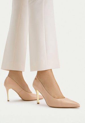 Kvinde iført beige spidse højhælede sko med gyldne stilethæle og hvide cropped vidde bukser på hvid baggrund.