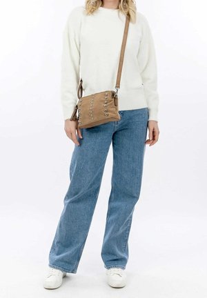 Sac bandoulière en daim marron clair avec des clous argentés, une sangle réglable et une fermeture éclair. Porté avec un pull blanc et un jean bleu.