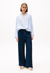 Blusa blu chiaro con scollatura a V e dettaglio arricciato all'apertura, abbinata a pantaloni a gamba larga blu navy. Si indossano sandali con cinturino beige.