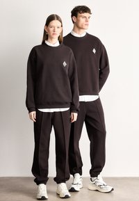 Σκούρες καφέ φούτερ και αντίστοιχα παντελόνια φόρμας με ελαστικές μανσέτες. Σημαντικό λευκό λογότυπο διαμαντιού στο στήθος. Καθημερινό αθλητικό στυλ.