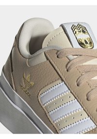 Lähenemine bež ja valge Adidas tossule kuldse logoga külgedel ja keeles, bež paelte ja tekstuuriga tallaga.