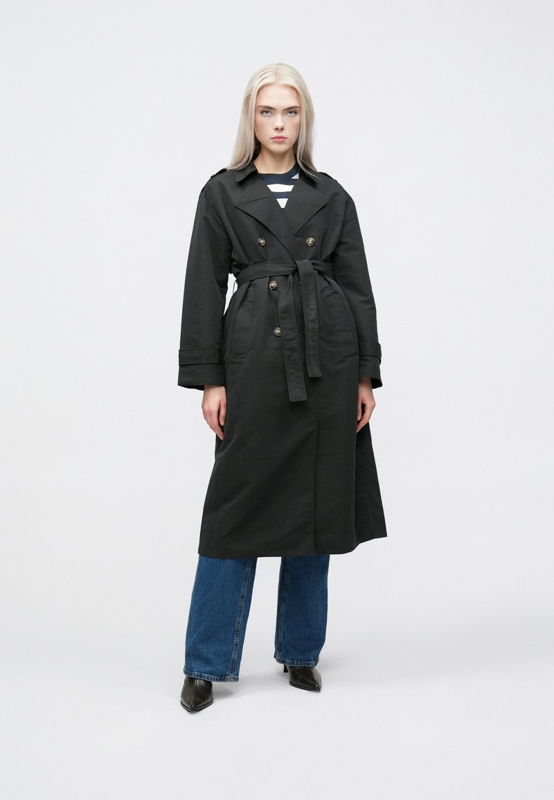 Cappotto nero lungo con vita regolabile, design doppiopetto, spalline sulle spalle e tasche frontali. Abbinato a jeans a vita alta blu a zampa d'elefante.