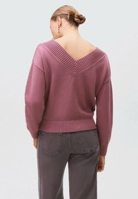 Rožnato pleteno pulover z širokim V-izrezom in rebrastimi detajli na izrezu in robu, v kombinaciji s sivoh hlačami.