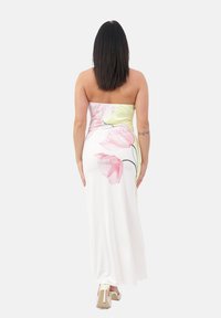 Femme aux cheveux sombres raides portant une robe blanche sans bretelles avec un motif floral rose et des chaussures à talons hauts blanches, de dos.