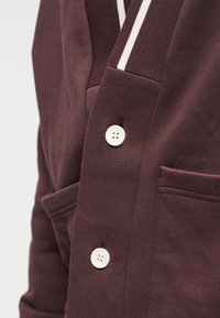 Pojem blizu temno rjave jopice z belimi gumbi in črtastim belim in rjavim ovratnikom, ki prikazuje teksturo in podrobnosti žepa.