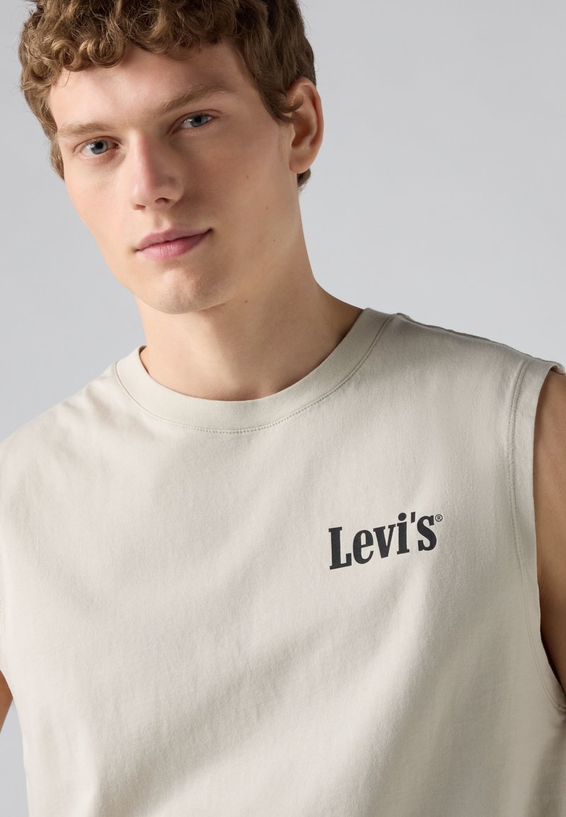 Jeune homme aux cheveux courts et bouclés portant un débardeur beige Levi's, regardant légèrement vers la caméra sur un fond uni.
