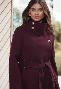 Burgundi gyapjú kabát magas gallérral, arany gombokkal és megkötős övvel. Strukturált kialakítással és sima textúrával rendelkezik.