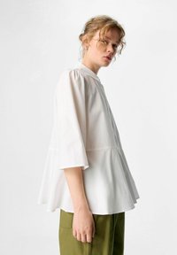 Weißes Peplum-Bluse mit Drei-Viertel-Ärmeln, einem runden Kragen und einem subtilen Glanz, kombiniert mit olivgrünen Hosen.