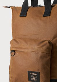 Barna viaszolt vászon hátizsák elülső zsebével, fekete hardverrel és márkázott folttal. Fekete állítható pántokkal és felső fogantyúval rendelkezik.