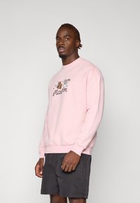 Felpa rosa in misto cotone con scollo rotondo e polsini a costine, con grafiche di cartoni animati e scritte sul davanti. Indossata con pantaloni corti scuri.