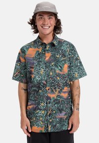 Camisa de manga corta con un colorido patrón de hojas tropicales, predominantemente verde con acentos naranjas, hecha de una tela suave y con botones.