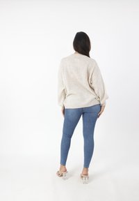 Beige Strickpullover mit überschnittenen Schultern, kombiniert mit blauen Skinny Jeans. Das Modell trägt transparente Absatzsandalen und ist von hinten vor einem weißen Hintergrund zu sehen.