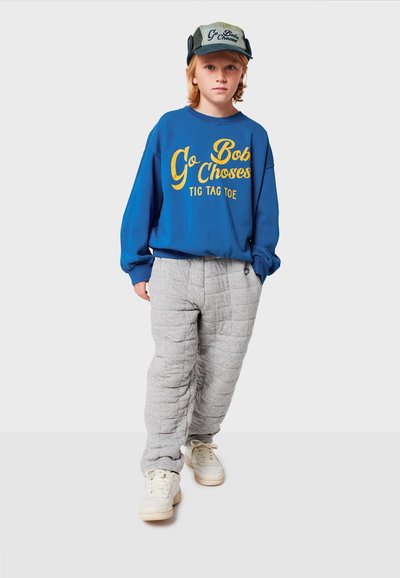 Felpa blu con testo e graphic gialli, pantaloni grigi trapuntati, sneakers bianche e un cappellino verde con testo ricamato.