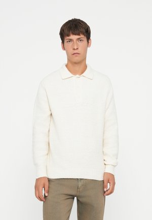 Cream-colored knitted sweater with a collared neckline, featuring a button placket and ribbed cuffs. Paired with light beige pants.