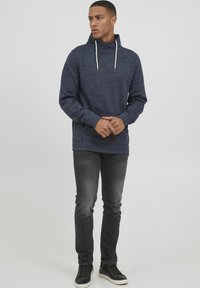 Sötétkék pulóver magas gallérral és húzózsinórral, textúrált anyagból készült. Sötétszürke farmerrel és fekete tornacipőkkel párosítva.