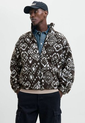 Chaqueta de forro polar marrón y blanca con un patrón, cuello alto, cierre de cremallera y puños elásticos; llevada sobre una camisa de mezclilla.