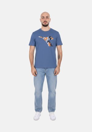 Uomo con barba e tatuaggio in piedi di fronte, indossa una t-shirt grafica blu, jeans azzurri e sneaker bianche su uno sfondo neutro.