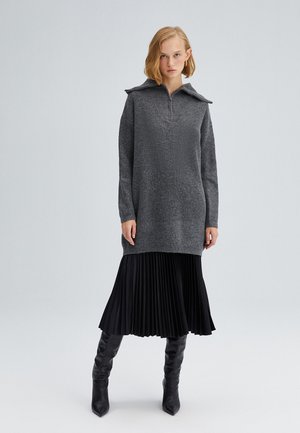 Grey knitted tunic with a collar and zip detail, layered over a black pleated skirt, paired with black knee-high boots.