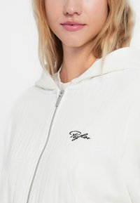 Weiße Sweatjacke mit Reißverschluss, geripptem Material, Kapuze mit Kordelzug und schwarzem gesticktem Logo auf der linken Seite.