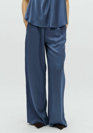 Osoba nosząca luźne, błyszczące, ciemnoniebieskie spodnie o szerokich nogawkach oraz dopasowaną bluzkę, stojąca z rękami w kieszeniach na jasnym tle.