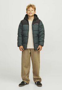 Verstevigde jas in donkergroen en zwart, met een rits, capuchon en logo op de borst. Geken hébt over een licht shirt en gecombineerd met beige broek.