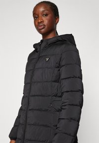 Doudoune noire avec capuche, présentant une texture matelassée et un logo triangulaire. Fermeture zippée et poches latérales.
