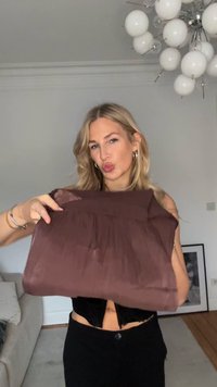 Brun, plisseret stofpose holdt af en person, med en glat tekstur og en rektangulær form. Designet er enkelt og minimalistisk.