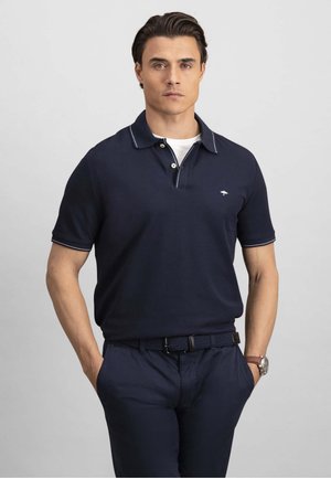 Mees, kes kannab tumesinist polot valge ääristusega ja tumesiniseid pükse, seisab käed taskutes ühetaolise heledahall tausta ees.