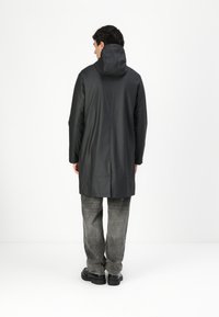 Long black raincoat with a hood, featuring a straight cut, smooth texture, and a back vent. Worn with gray jeans and black shoes.