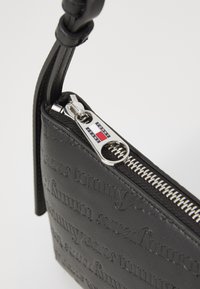 Black leather zipper pouch with embossed "Tommy Jeans" text, silver zipper hardware, and a braided strap detail.