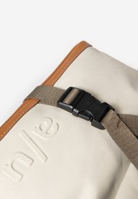 Beige canvas tas met beige leren accenten, voorzien van een zwarte kunststof gespsluiting en een getextureerd logodetail. De band is donkergrijs.