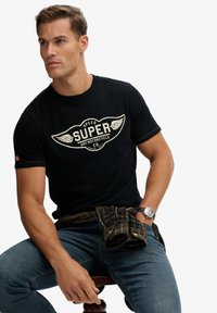 Camiseta negra de algodón con un gráfico crema que muestra alas y el texto "SUPER". Combinada con vaqueros azules y una camisa de franela a cuadros atada en la cintura.