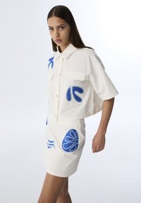 Weißes, gekürztes Shirt mit kurzen Ärmeln und blauen, gestickten Designs, kombiniert mit einem passenden, taillierten Rock, der ähnliche Akzente aufweist.