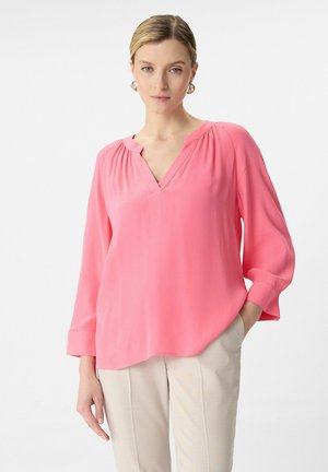 Rosa Langarmbluse mit V-Ausschnitt, gerafften Details an der Schulter, glatter Textur und lässigem Schnitt. Kombiniert mit hellen Hosen.