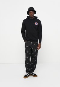 Zwarte hoodie met een voorzak en logo, gecombineerd met zwarte jeans met een yin-yang patroon, afgewerkt met zwarte sneakers en een bucket hat.