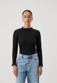 Black long-sleeve top with ribbed texture and a ruffled neckline, paired with light blue high-waisted jeans featuring button details.
