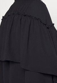 Schwarzes Kleidungsstück mit geschichtetem Stoff, das Rüschenakzente entlang der Nähte aufweist. Weicher Griff, der elegant über die Schultern fällt.