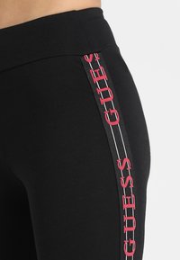 Leggings neri con una texture liscia, dotati di un prominente logo rosso "GUESS" lungo il lato, bordato da strisce bianche.