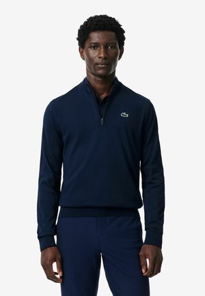 Uomo che indossa un maglione blu navy con zip a un quarto e piccolo logo verde di coccodrillo sul petto e pantaloni blu navy abbinati.